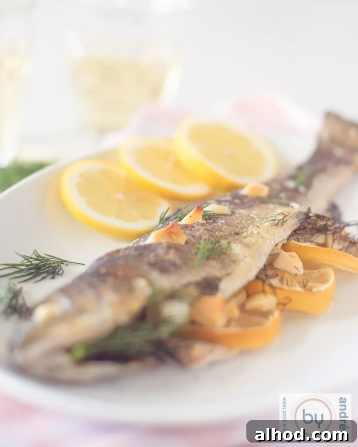 Zesty Lemon Dill Baked Trout 2 An eye-level photo of a white dish with a beautifully baked salmon trout, garnished with fresh lemon slices, vibrant dill sprigs, and thinly sliced garlic.