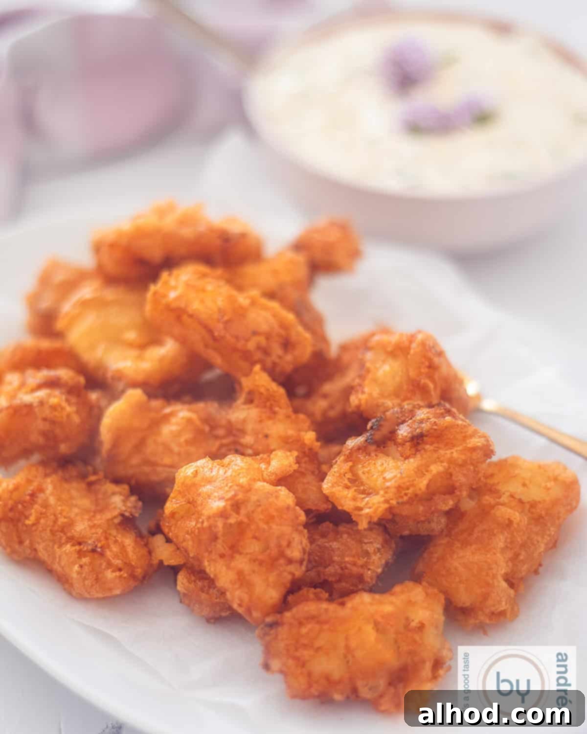 Golden Kibbeling Bites – Holland's Favorite Fried Fish 2 A height photo of a portion of kibbeling on a white plate. Some ravigotte sauce in the background