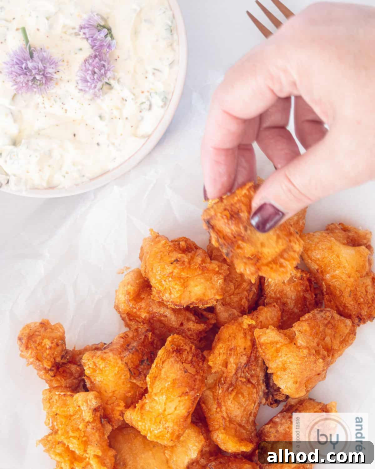 Golden Kibbeling Bites – Holland's Favorite Fried Fish 3 A height photo from above of kibbeling on a white plate. A hand grabs a piece of fish. Ravigotte sauce at the top left.