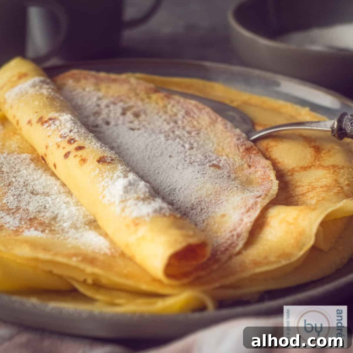 Simple Dutch Pannenkoeken from Scratch 4 A square photo with a dark gray plate with a stack of freshly baked pancakes with powdered sugar.