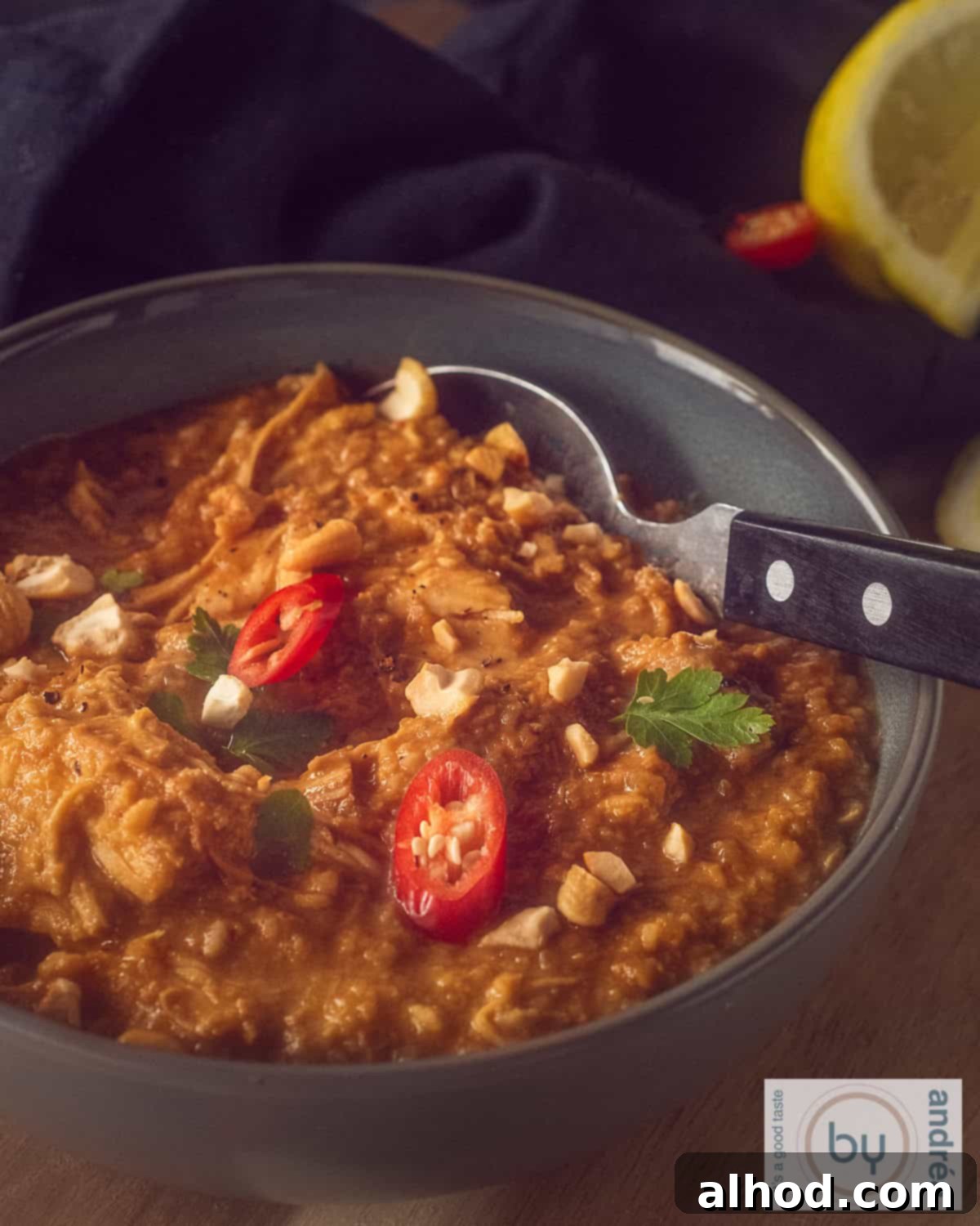 Creamy Slow Cooker Peanut Satay Chicken 2 A grey bowl with a spoon containing peanut sauce with chicken pieces. Garnish of red pepper and parsley and nuts.