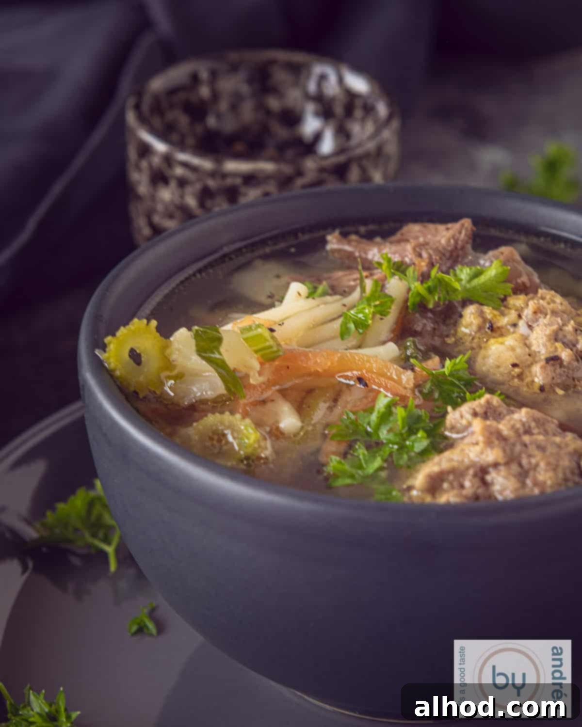 Hearty Dutch Meatball Vegetable Soup 3 A height photo of part of a gray bowl of vegetable soup with meatballs and parsley on a dark background.