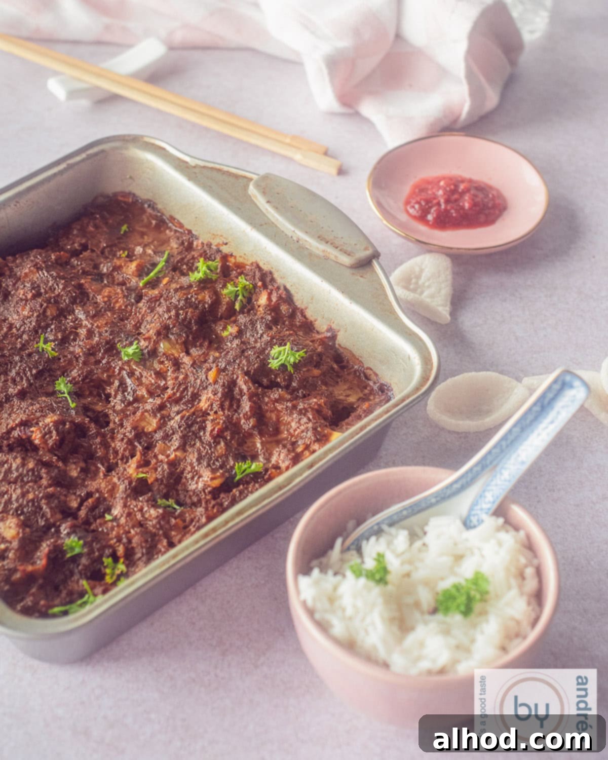 An eye-level photo featuring a rustic baking dish filled with Indonesian Frikadel Pan, surrounded by a small bowl of steamed white rice and a vibrant pink plate of sambal, showcasing a complete and inviting meal.
