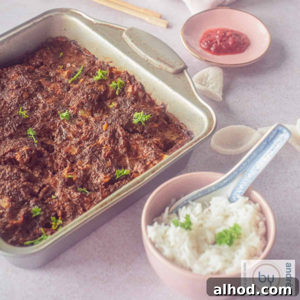 A square photo of a rectangular baking dish filled with the uncooked Indonesian Frikadel Pan mixture, ready for the oven. Beside it, a pink bowl holds steamed white rice and a small dish contains vibrant sambal, hinting at the complete meal.