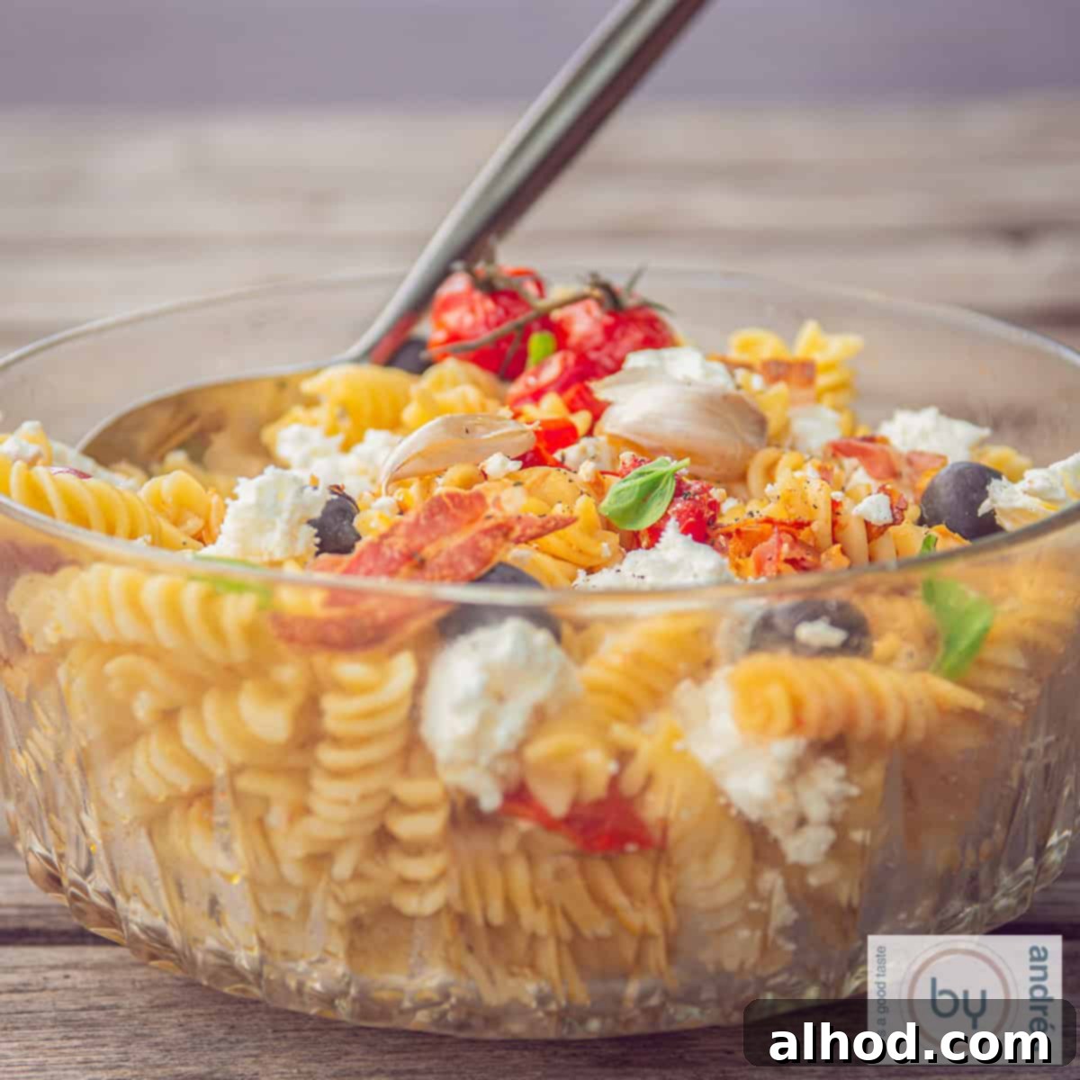 Mediterranean Bacon Feta Pasta Salad 4 A square photo with a glass bowl on a wooden background filled with pasta salad, tomatoes, bacon and feta