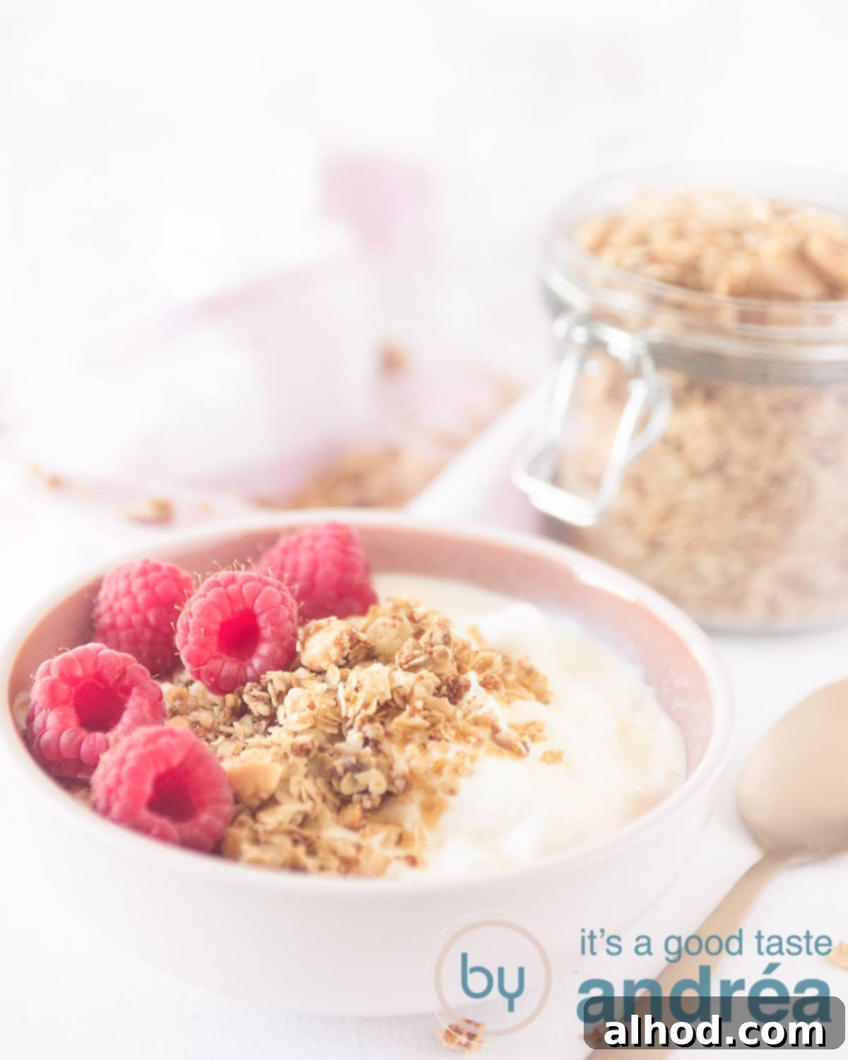 Homemade Almond Coconut Granola Made Simple 2 A pink bowl filled with creamy yogurt, homemade almond coconut granola, and fresh raspberries. In the background, a glass jar brimmed with more golden granola and a golden spoon rests on the right.
