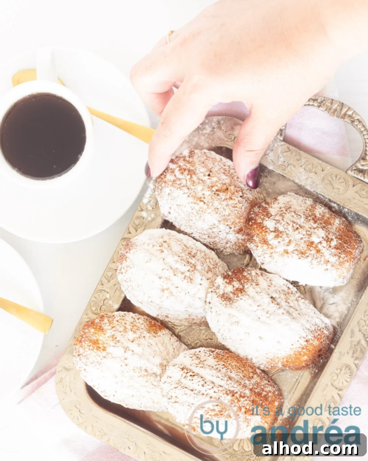 A hand taking a madeleine from a bowl of six madeleines. A cup of coffee on the background