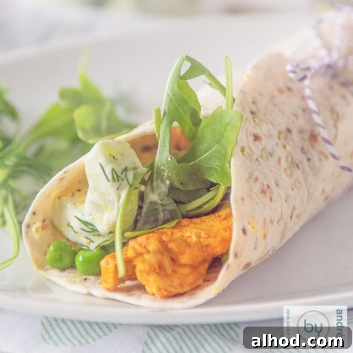 A square photo of a white plate with a rolled wrap filled with Tandoori chicken, tzatziki and fresh vegetables, garnished and ready to eat.