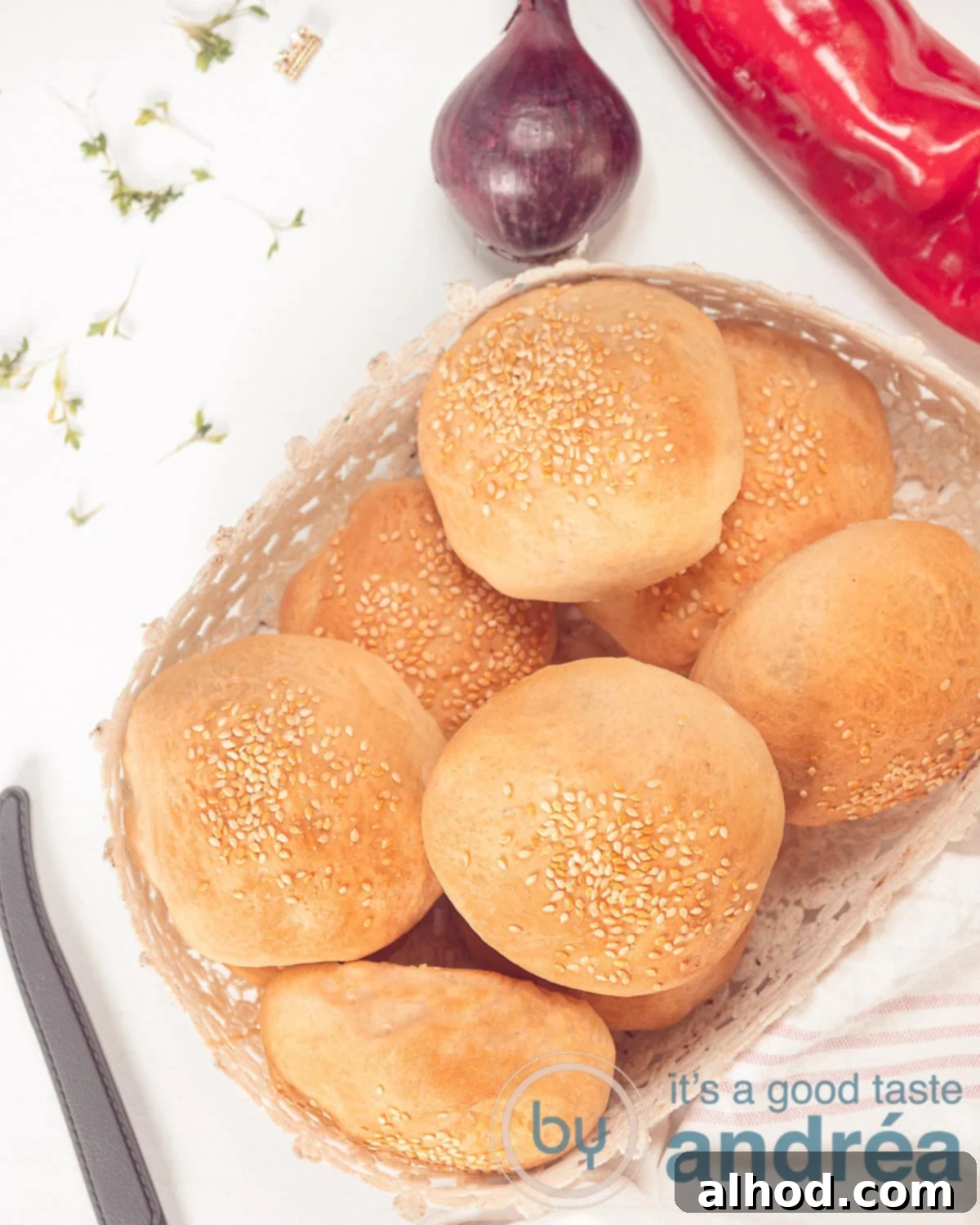 A photo from above of a white bowl filled with fresh hamburger buns. Top right a fresh onion and bell pepper, top left cress.