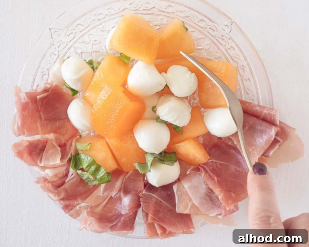 Vibrant Melon & Prosciutto Salad with Creamy Mozzarella 6 A glass plate with parmaham, mozzarella, melon, basil and dressing. A fork mixing everything.