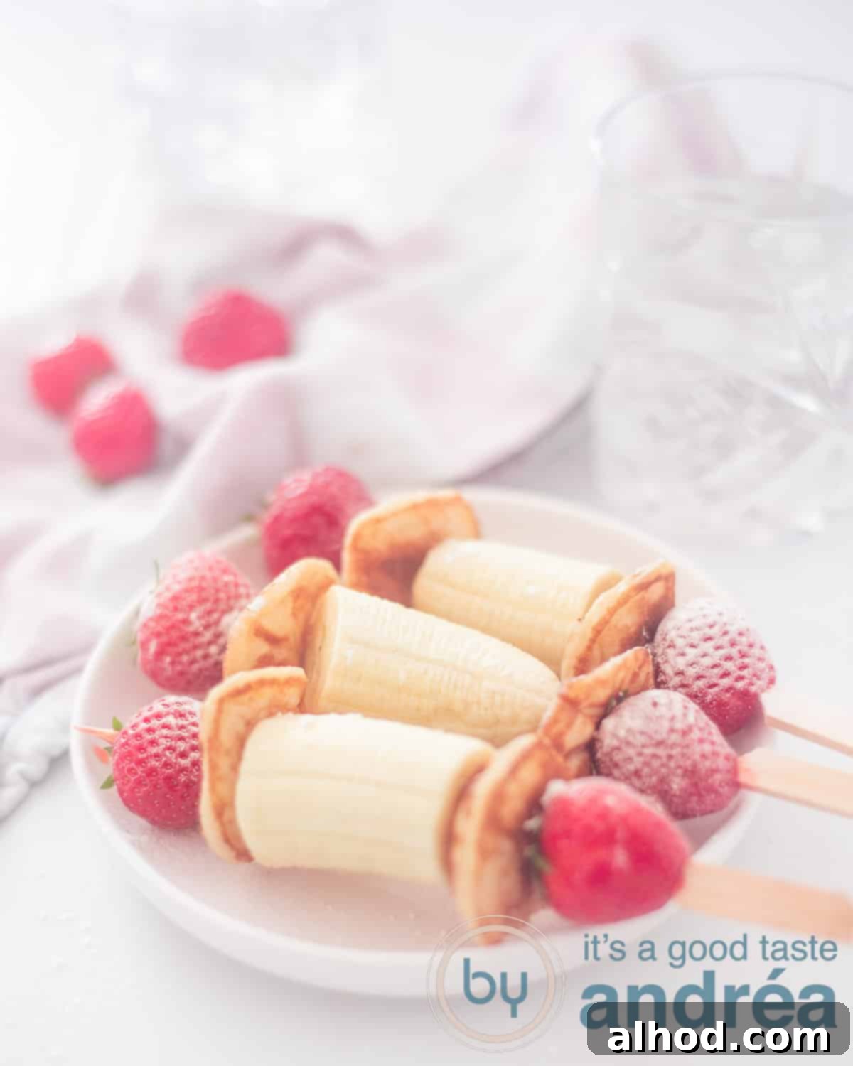 Strawberry Banana Pancake Pops 2 An elevation photo of a white plate with three skewers with strawberry, banana and pancakes. In the background glasses and strawberries. Everything on a white background