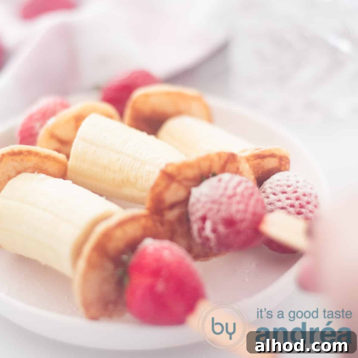 A square photo displaying three delightful skewers, each alternating with slices of banana, fluffy pancakes, and fresh strawberries. A hand is reaching for one from the board.