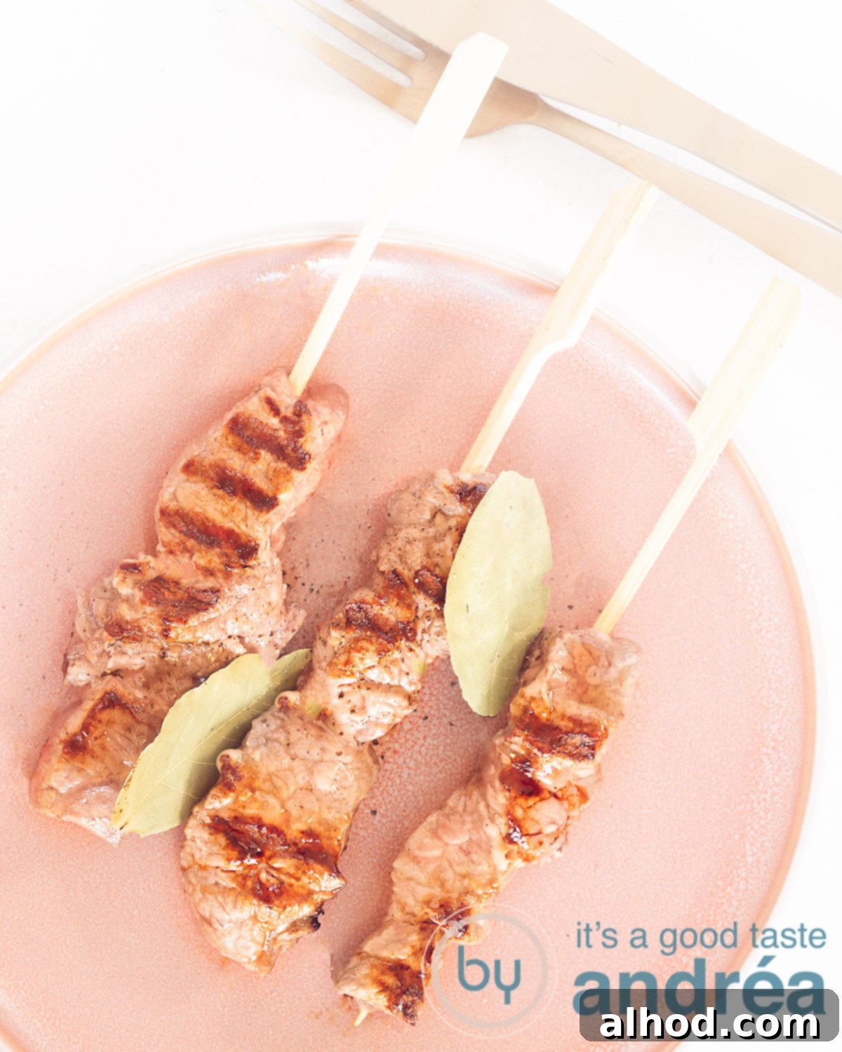 Madeira Style Beef Skewers 2 A photo from above of a pink plate with espetadas and bay leaves on a white background, ready to be served.