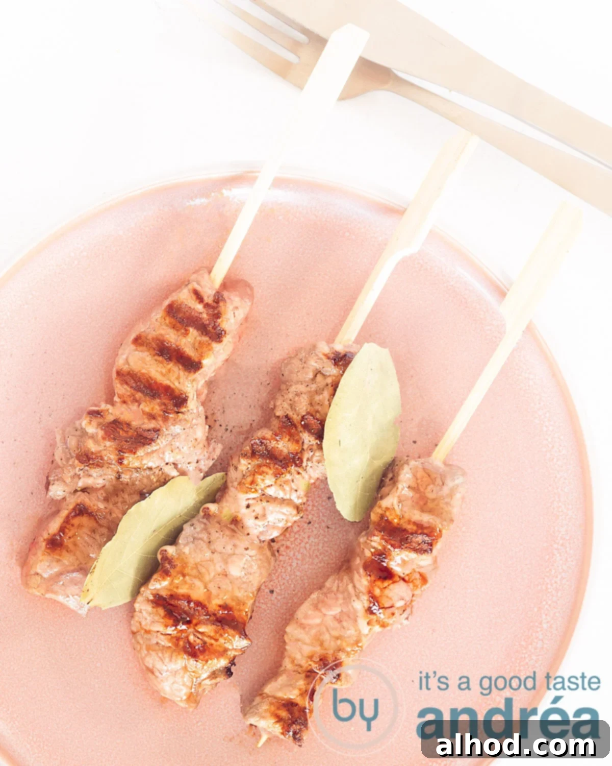 Espetada Madeira Beef Skewers 2 A photo from above of a pink plate with espetadas and bay leaves on a white background. Delicious grilled Portuguese beef skewers.