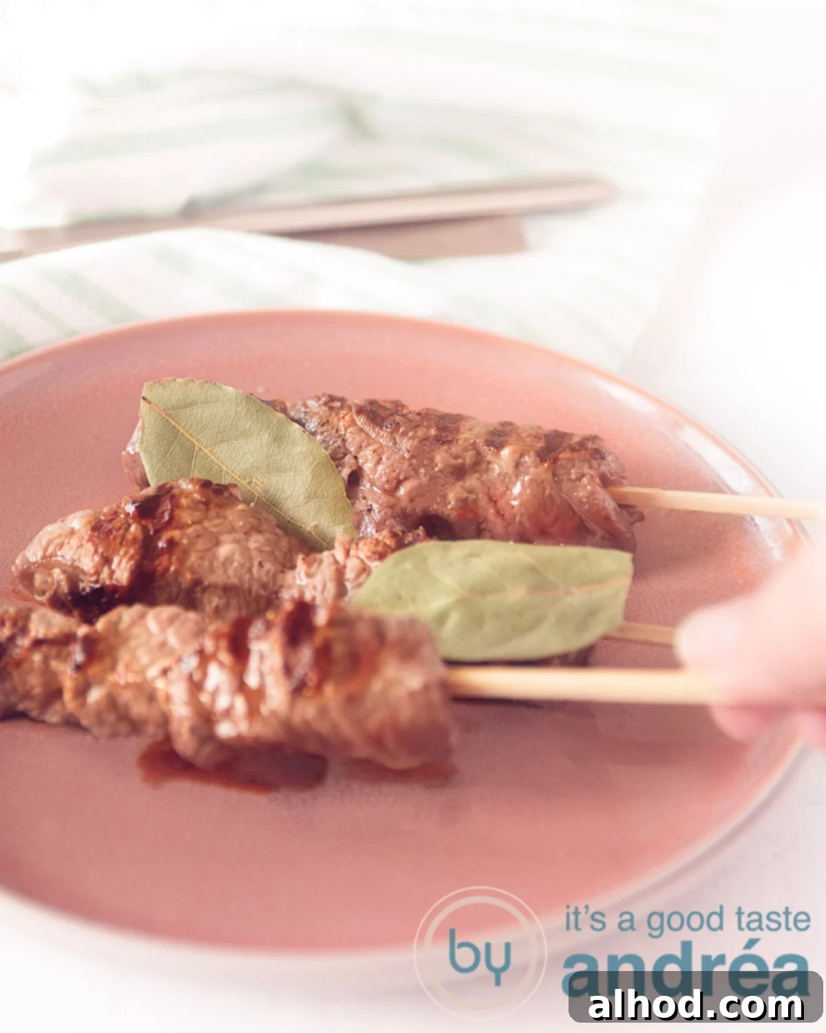 Madeira Style Beef Skewers 5 Three Portuguese sirloin skewers on a pink plate with bay leaves. A hand is reaching for a skewer, showing the tender, grilled meat.