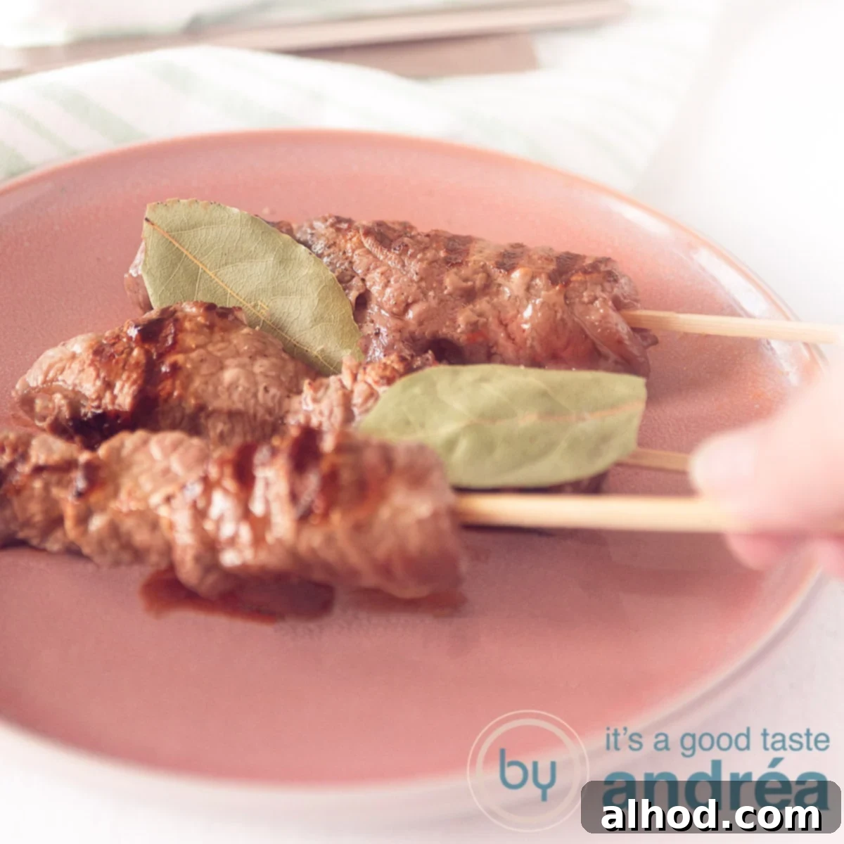 Madeira Style Beef Skewers 6 A square photo with a pink plate on which three Portuguese grilled skewers. A hand takes a skewer