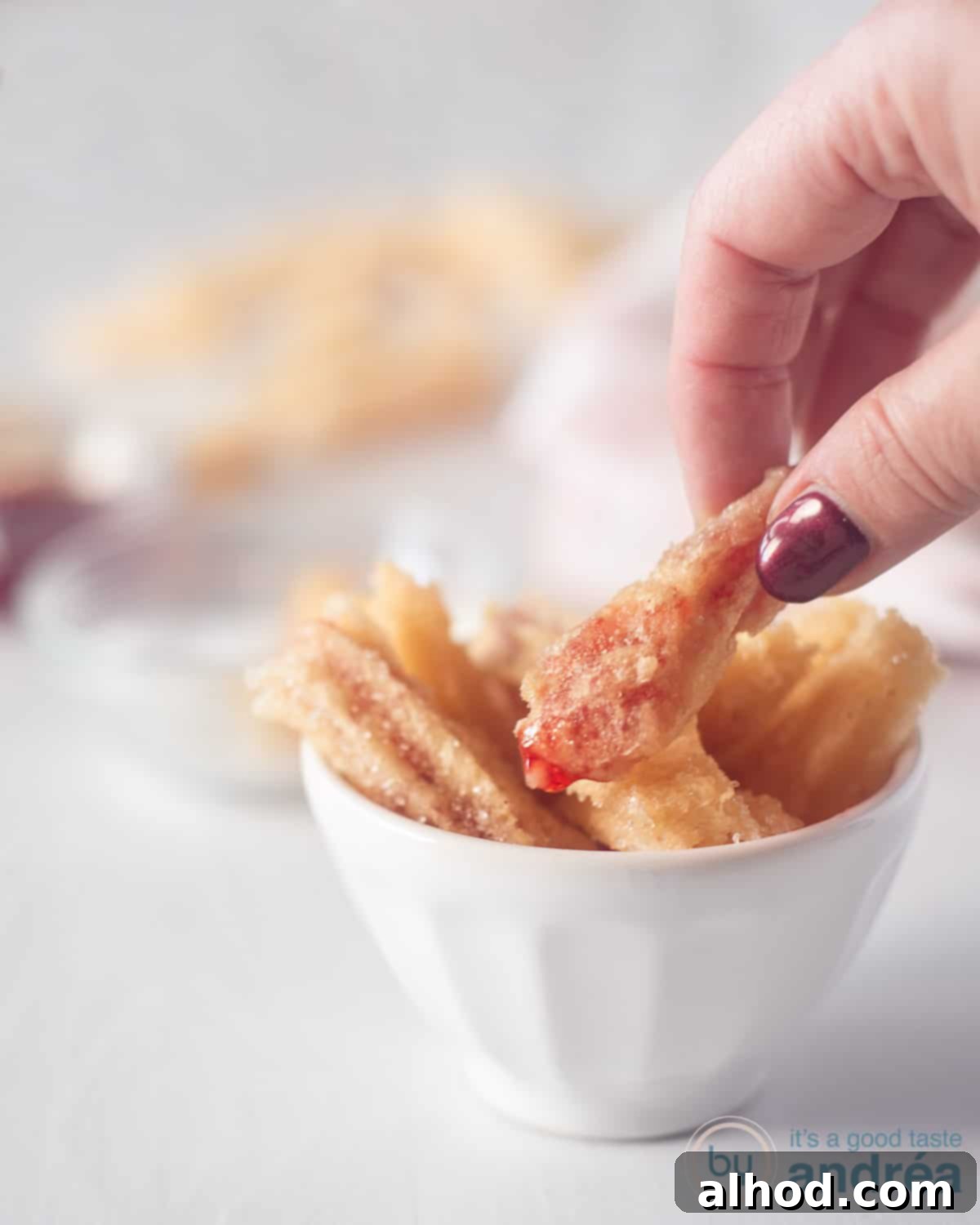 Strawberry Burst Churros 3 A height photo with a white container filled with Churros filled with strawberry jam. A hand takes a churro from the container.