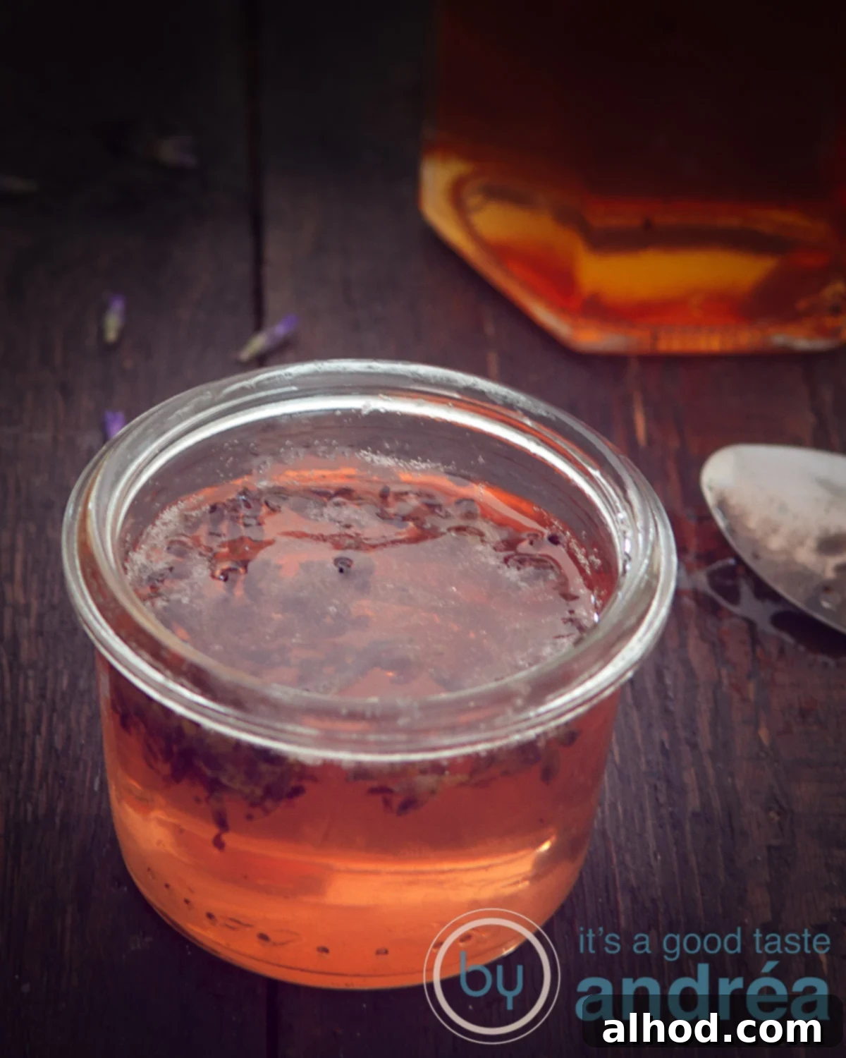 Lavender Kissed Apple Jelly 2 A macro photo of a jar filled with shimmering caramel-colored apple and lavender jelly, set against a dark background, highlighting its smooth texture and rich hue.