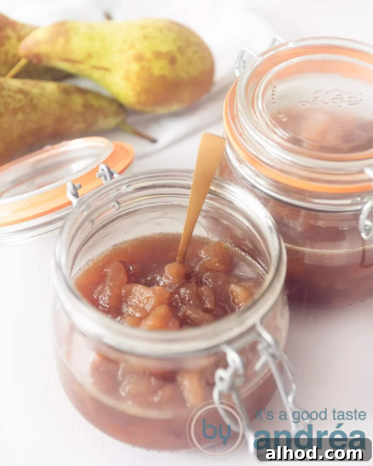 Warm Pear & Spice Preserve 2 An open glass jar of pear jam with a golden spoon. A second pot diagonally behind it. Two pears in the back left