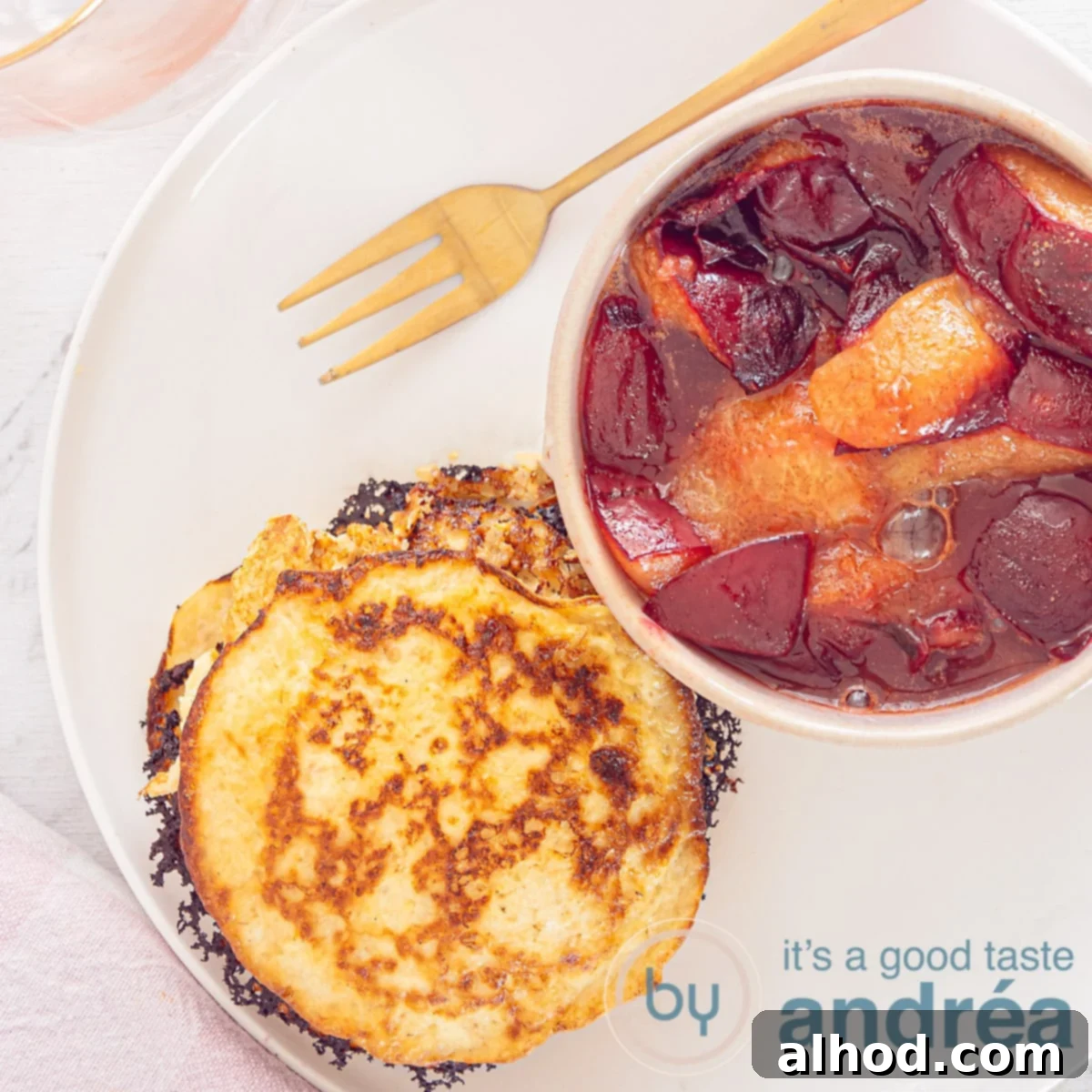 A square photo with pancakes and plum compote in a pink dish on a white plate. A golden fork lies above it.