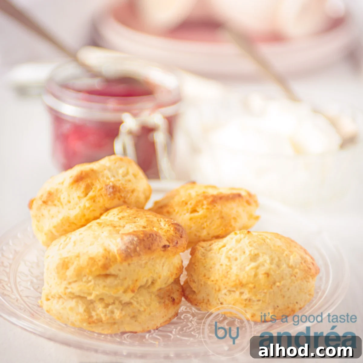 A square photo with a glass plate of scones and a jar of strawberry jam in the background.