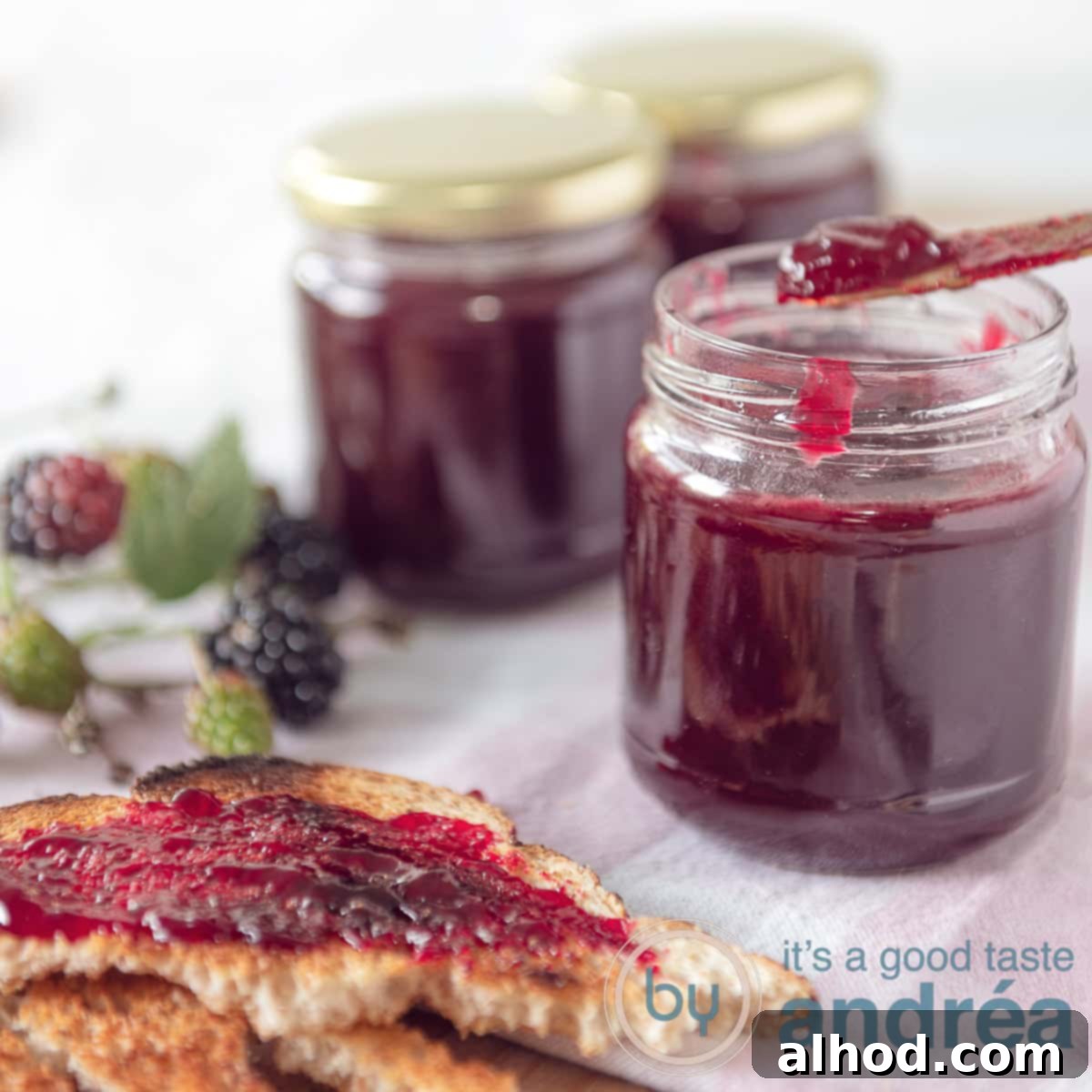 A square photo with three jars of blackberry jam with 1 opened from which a little is taken. In the front a slice of bread spread with jam.