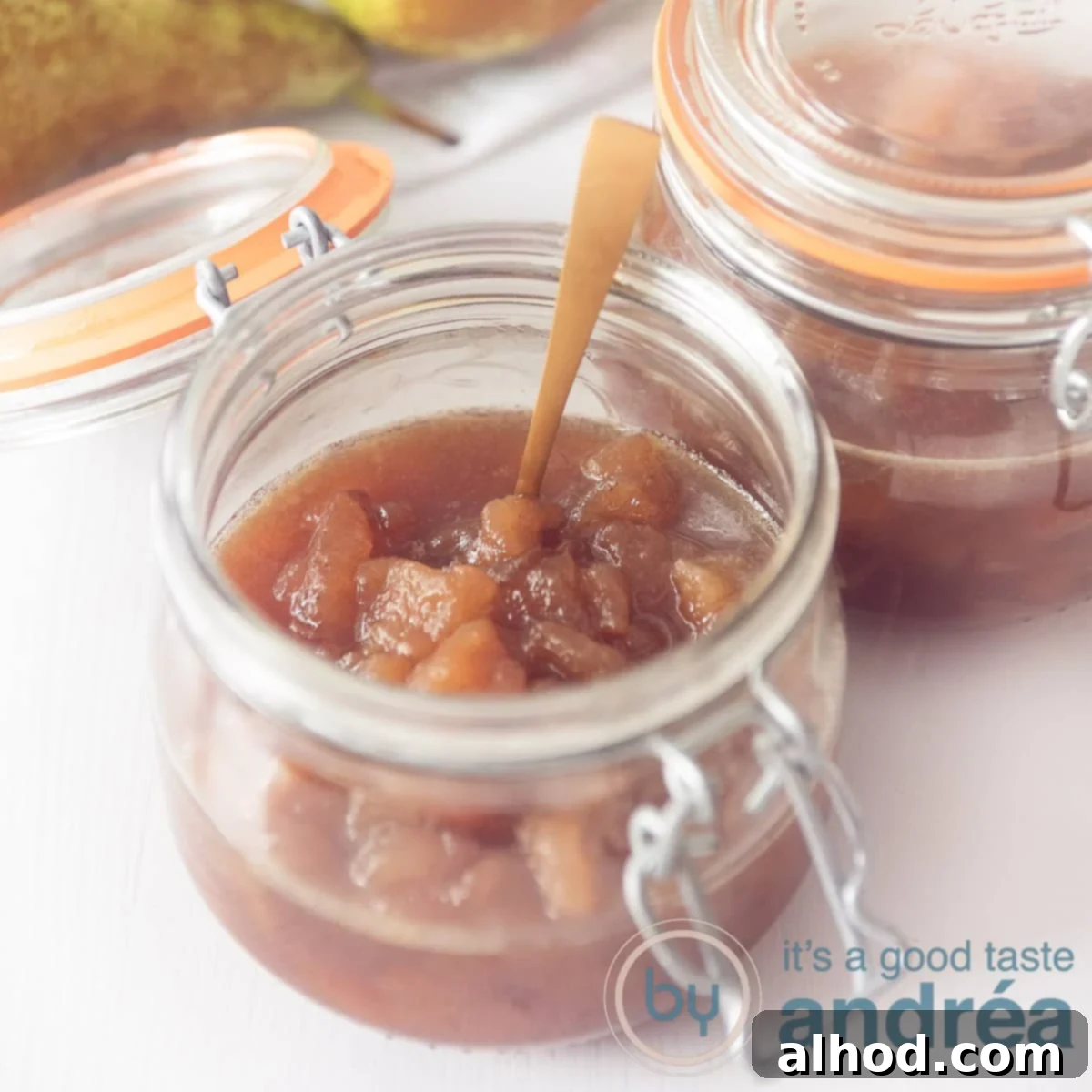 A square photo with an open jar of pear jam. A golden spoon in it on a white background