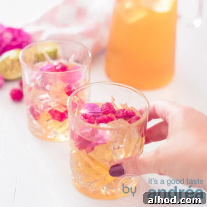 A square picture featuring two glasses and a jar of lime rose and raspberry iced tea arranged diagonally. A hand reaches for the first glass, indicating immediate enjoyment.