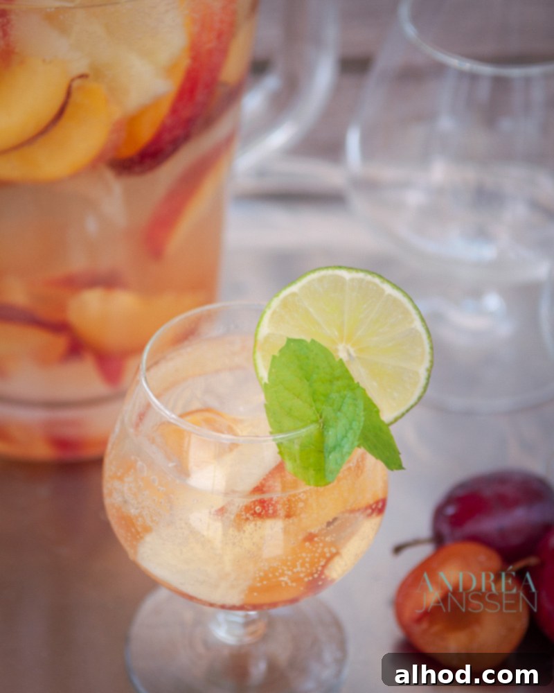 A vibrant glass of Sangria Bianca, generously filled with fresh fruit, and garnished with a sprig of mint and a lemon slice. A pitcher and more glasses are visible in the background, suggesting a festive setting.