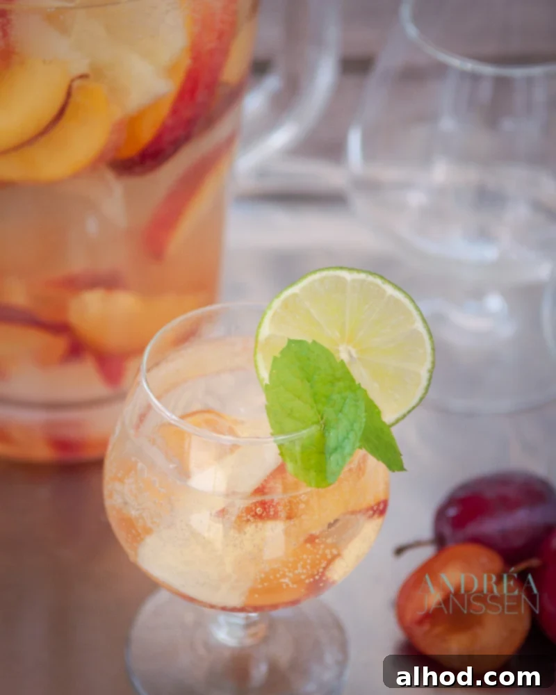 A glass of refreshing Sangria Bianca with an abundance of fresh fruit, garnished with mint and lemon, with a pitcher and other glasses in the background.