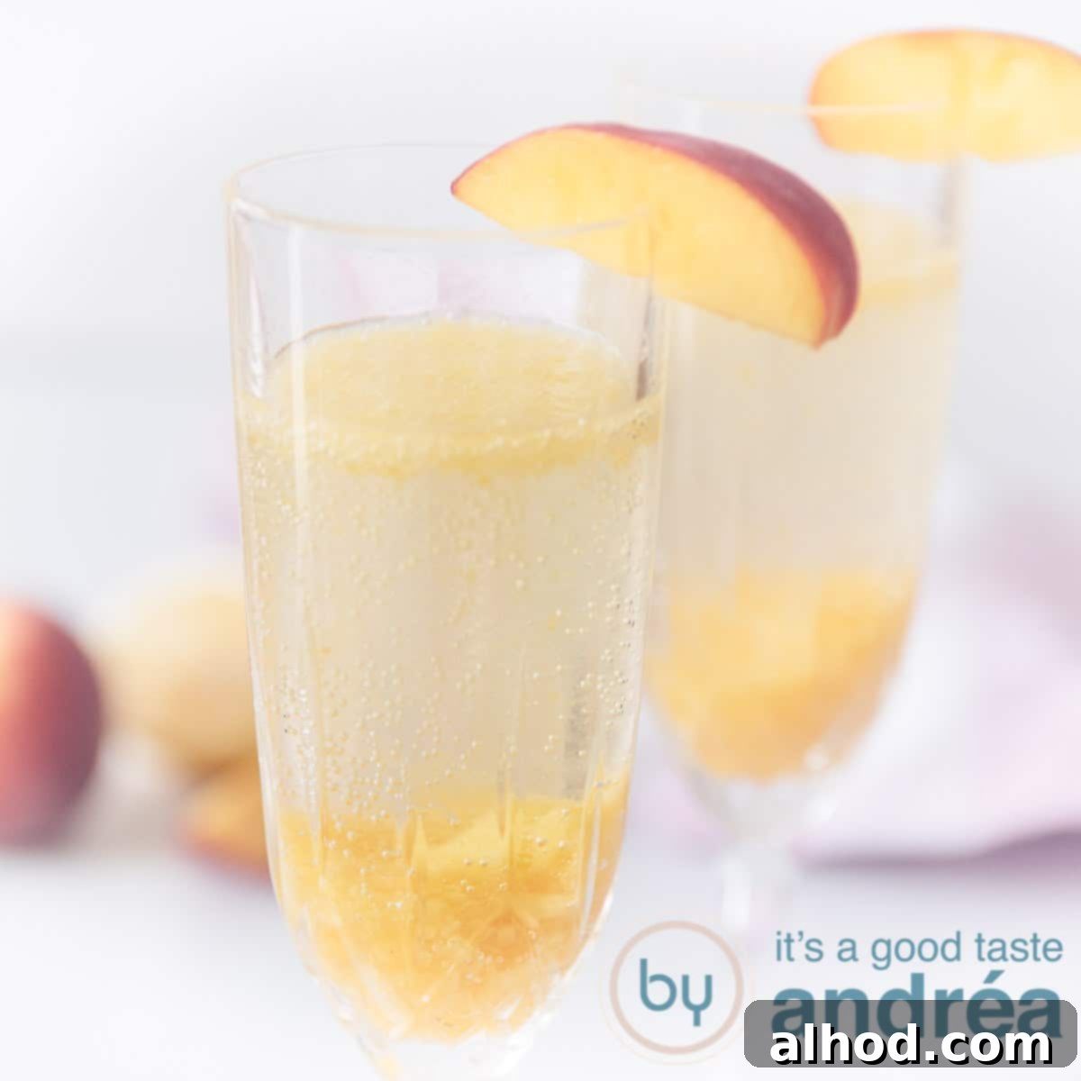 A square photo with two champagne glasses filled with a vibrant Bellini cocktail, with fresh peaches blurred in the background.