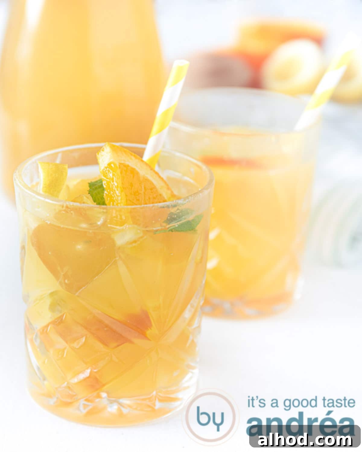Lemon Mint Cooler 2 Two glasses filled with vibrant orange, lemon, and mint iced tea, with a glass pitcher and fresh oranges in the background, set on a bright wooden surface.