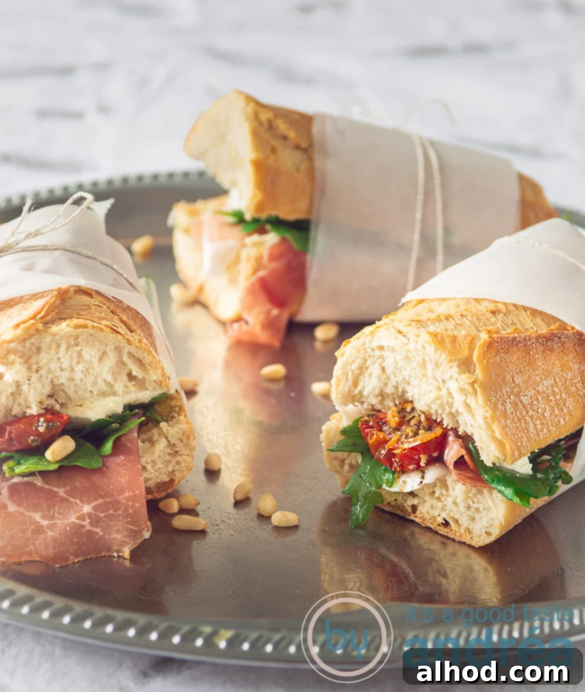 Parma Prosciutto Club 2 Three perfectly wrapped Italian Club Sandwiches with Prosciutto, Mozzarella, pesto, sun-dried tomatoes, and arugula on a silver serving plate, ready for a picnic or elegant lunch.