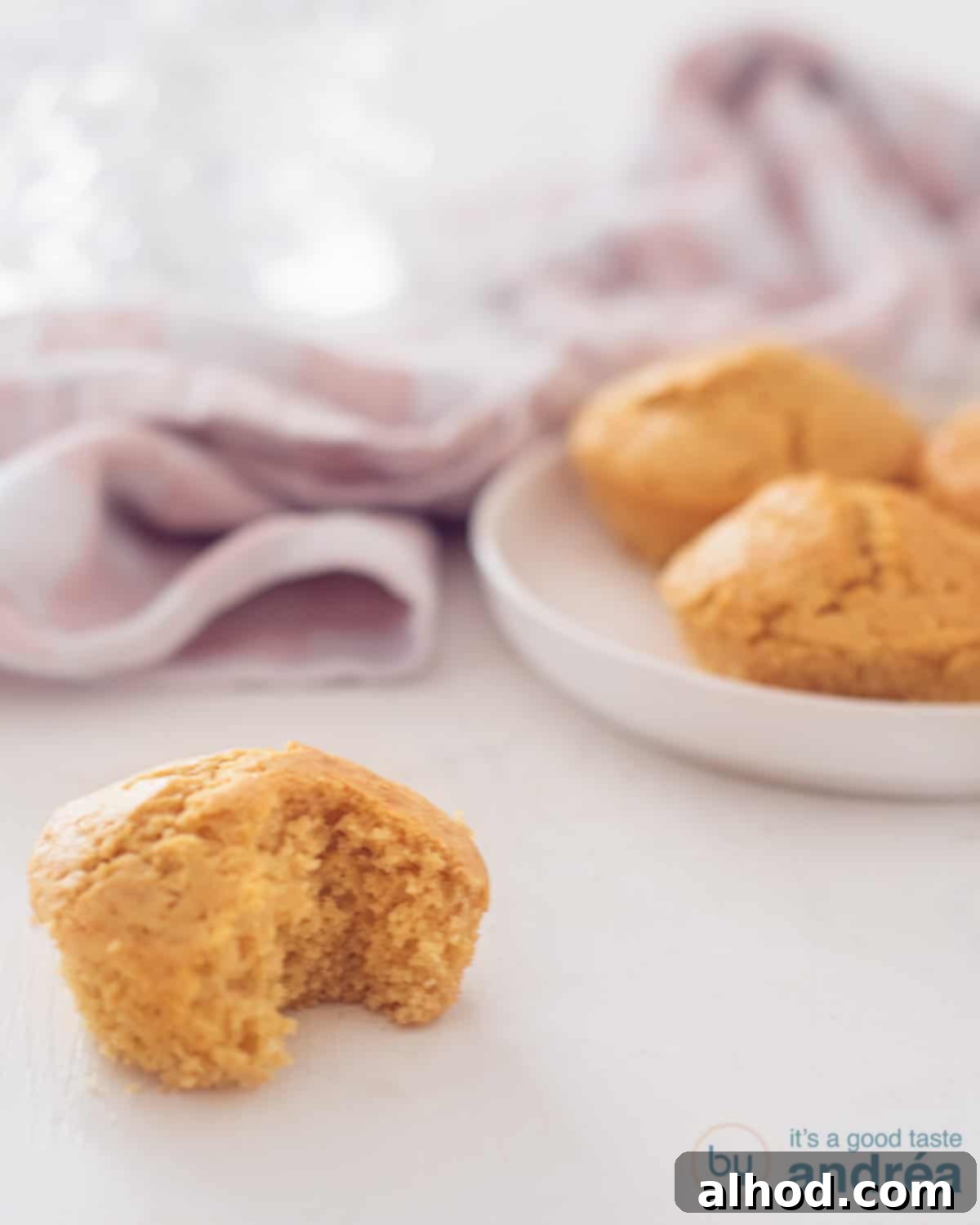 A perfectly baked corn muffin with a bite taken out, showcasing its fluffy interior, set on a white surface. Other golden muffins are blurred in the background, inviting a warm, homemade feel.