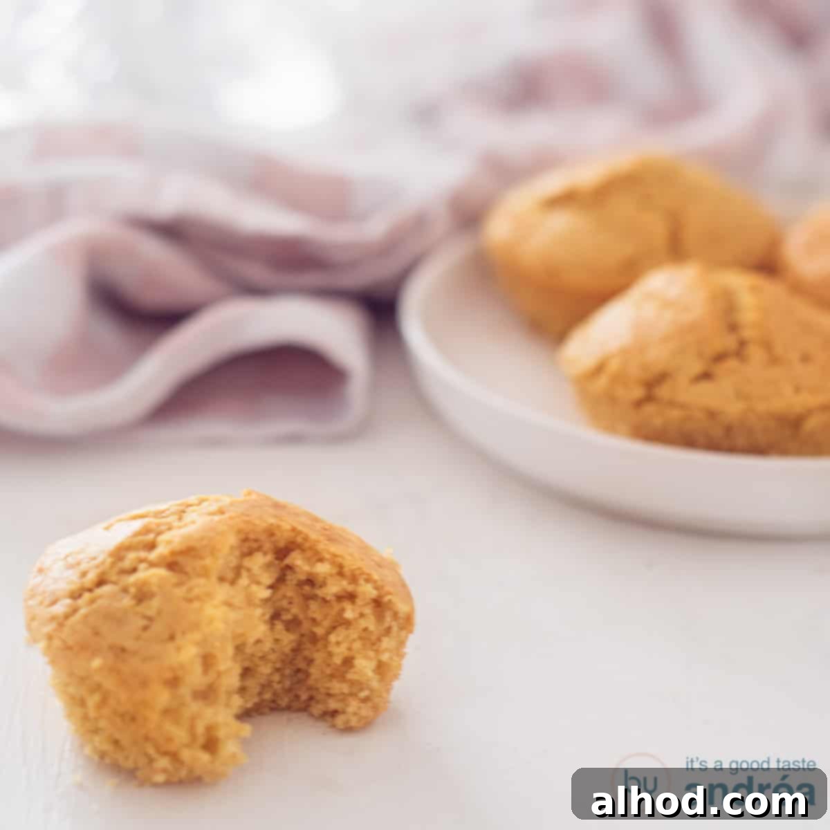 Golden Fluffy Cornbread Muffins Made Simple 4 A square photo of a single golden cornbread muffin with a bite taken, revealing its soft, airy texture, with a plate of other muffins in the background.