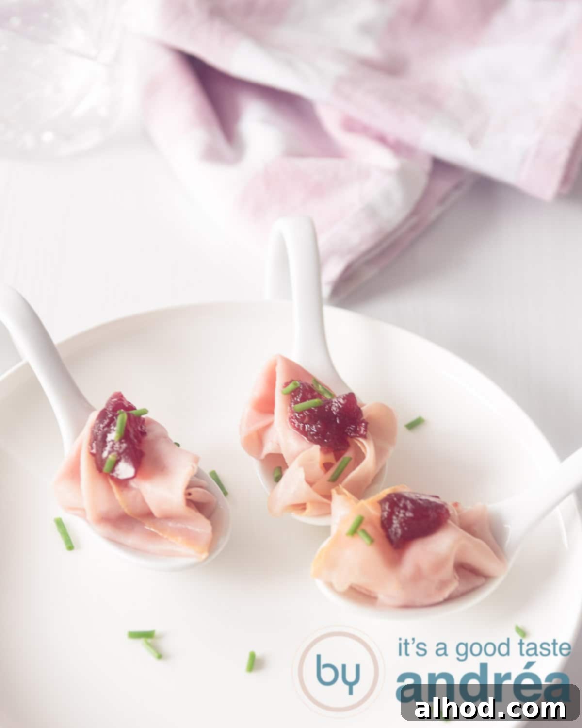 A pristine white plate showcases three elegant appetizer spoons, each holding a perfectly crafted portion of ham and cranberry amuse-bouche, set against a clean white background.