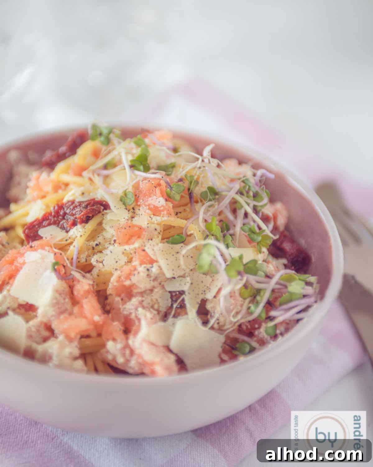 Velvety Smoked Salmon Pasta 3 A beautifully composed pink bowl, showcasing a swirl of creamy spaghetti adorned with tender flakes of smoked salmon, vibrant sun-dried tomato slices, and a dusting of grated Parmesan cheese. The rich sauce glistens under the light.