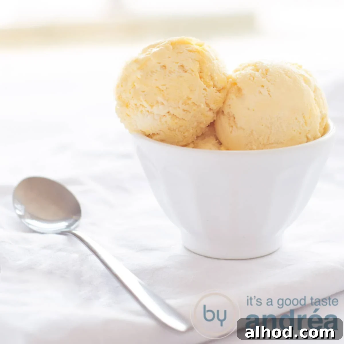 Decadent Cognac Ice Cream 2 A high-angle photo of a white bowl filled with scoops of golden-hued cognac ice cream. The bowl rests on a pristine white background, with a silver spoon placed to the left.