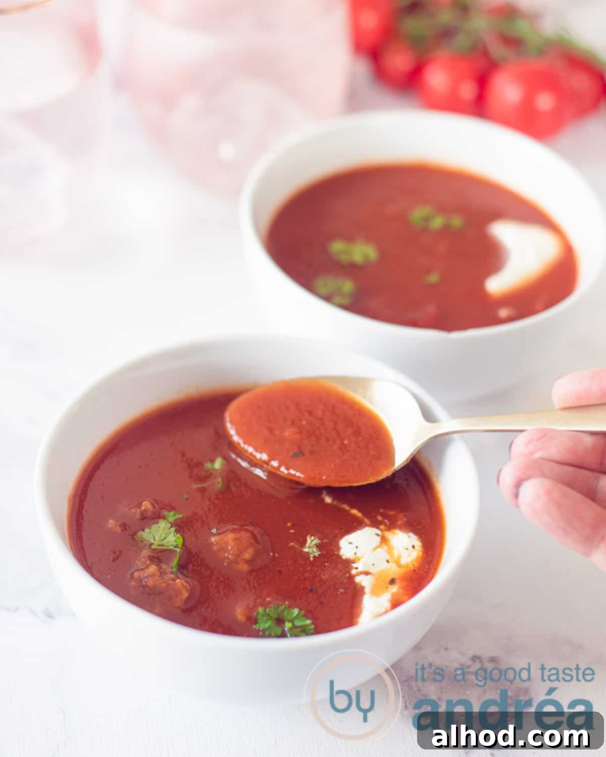Authentic Dutch Tomato Meatball Soup 3 Two white bowls filled with tomato soup with meatballs and a dollop of cream. A hand with a spoon taking a bite.