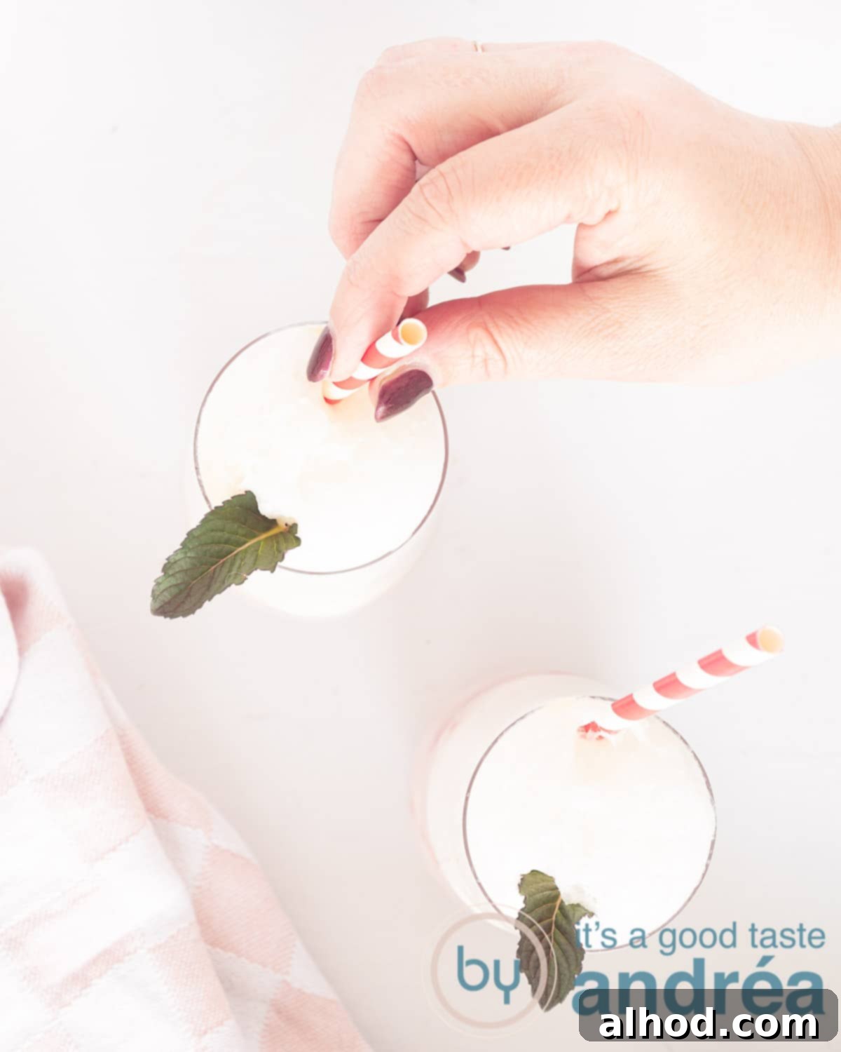 Strawberry Soda Float 2 A photo from above of two glasses with a foam layer and a mint leaf. A hand grabs a straw. A white background, showing the refreshing Strawberry Ice Cream Soda Float.