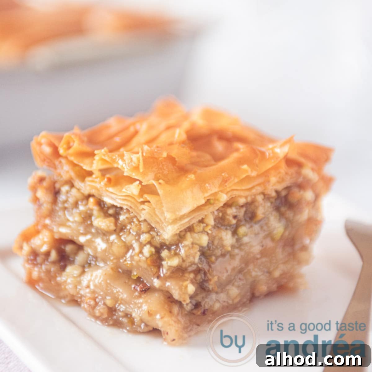 A square photo of a piece of baklava on a white plate. In the background a baking pan with the rest of the baklava