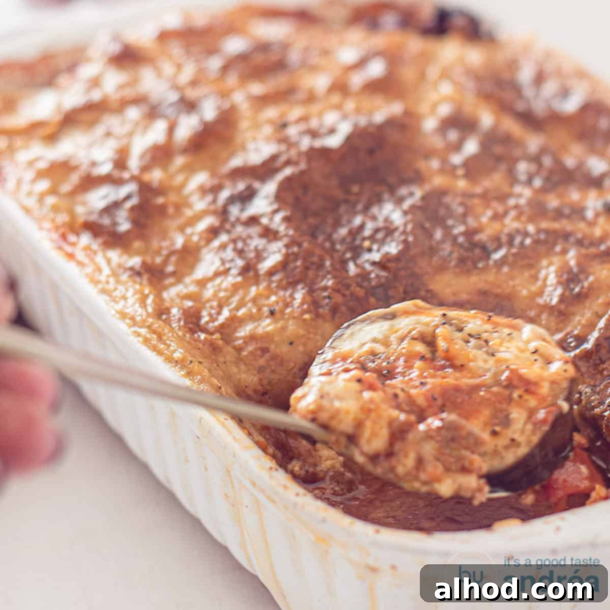 A square picture of a white casserole filled with Eggplant Moussaka. A spoon taking out some food.