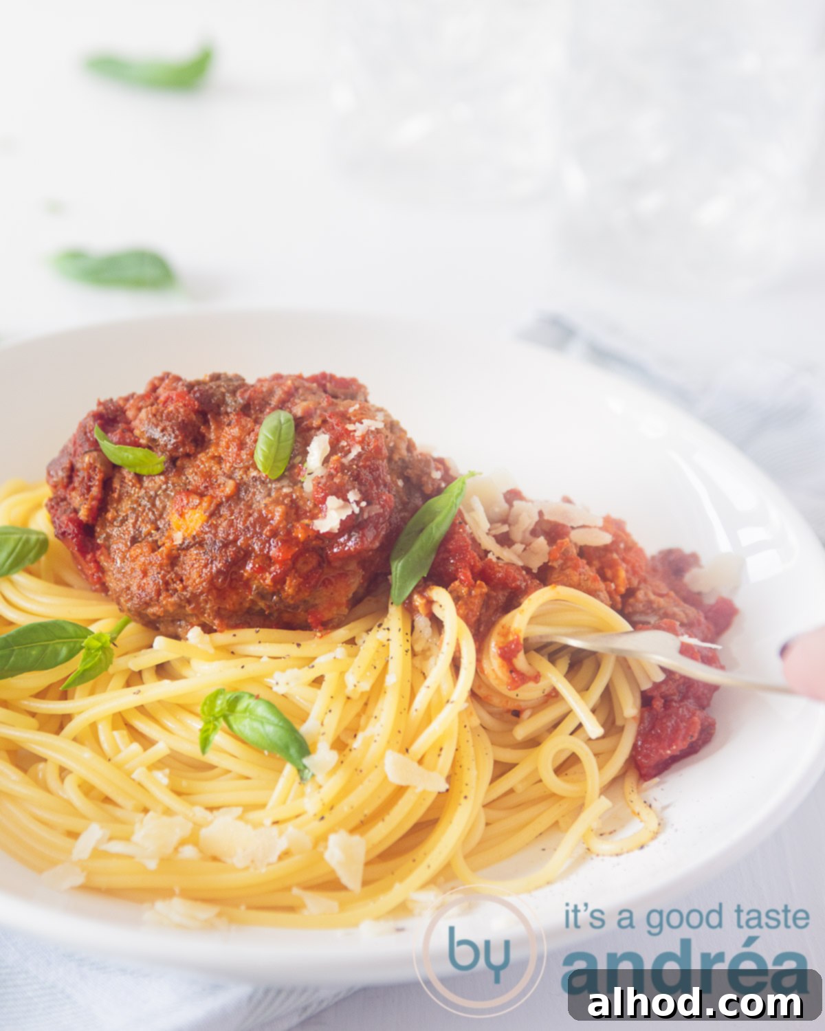Sage Meatball Skillet Parm 5 Meatballs with sage and parmesan cheese on a white plate with tomato sauce and spaghetti. A golden fork lies in it. A white background with glasses and basil leaves