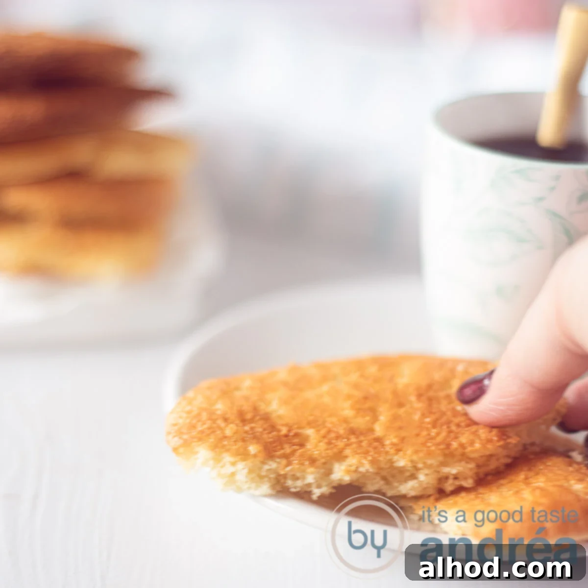 A pristine white plate showcases two halved Eierkoeken, revealing their airy interior, as a hand reaches to pick up another. A stack of these classic Dutch sponge cookies is visible in the background, ready to be enjoyed.
