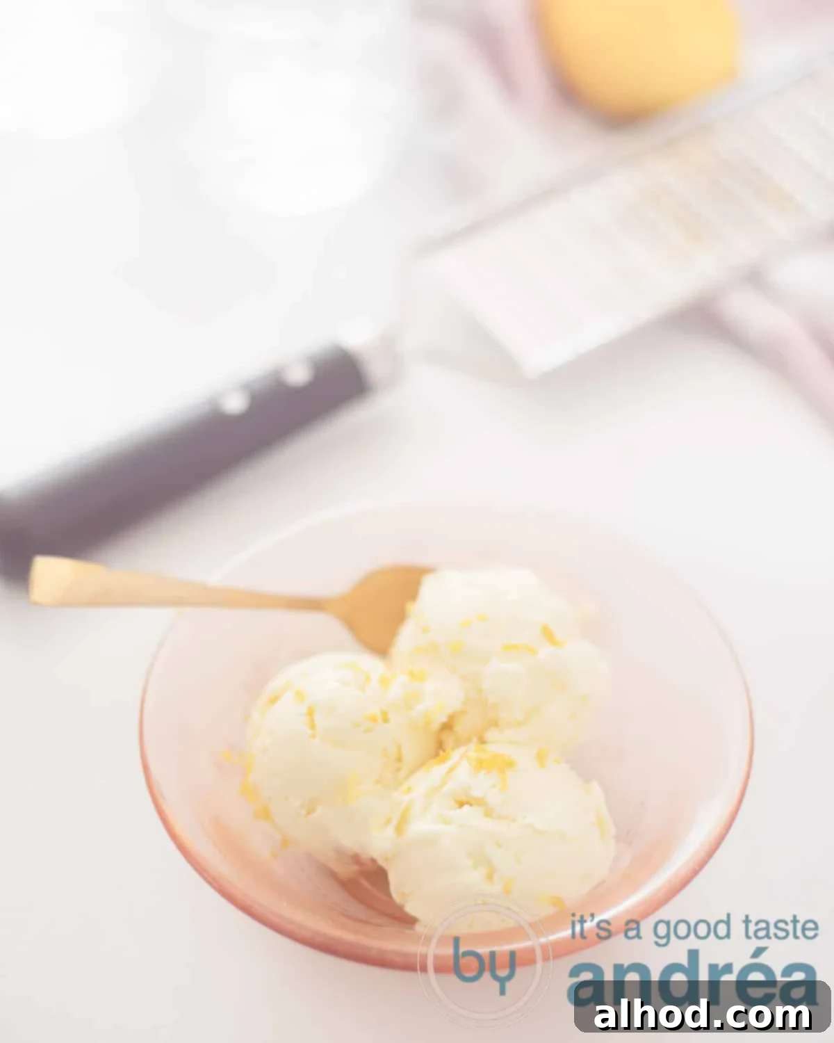 An elevation photo with an orange bowl with three scoops of lemon mascarpone ice cream with lemon zest. A grater and fresh lemon are visible in the background.