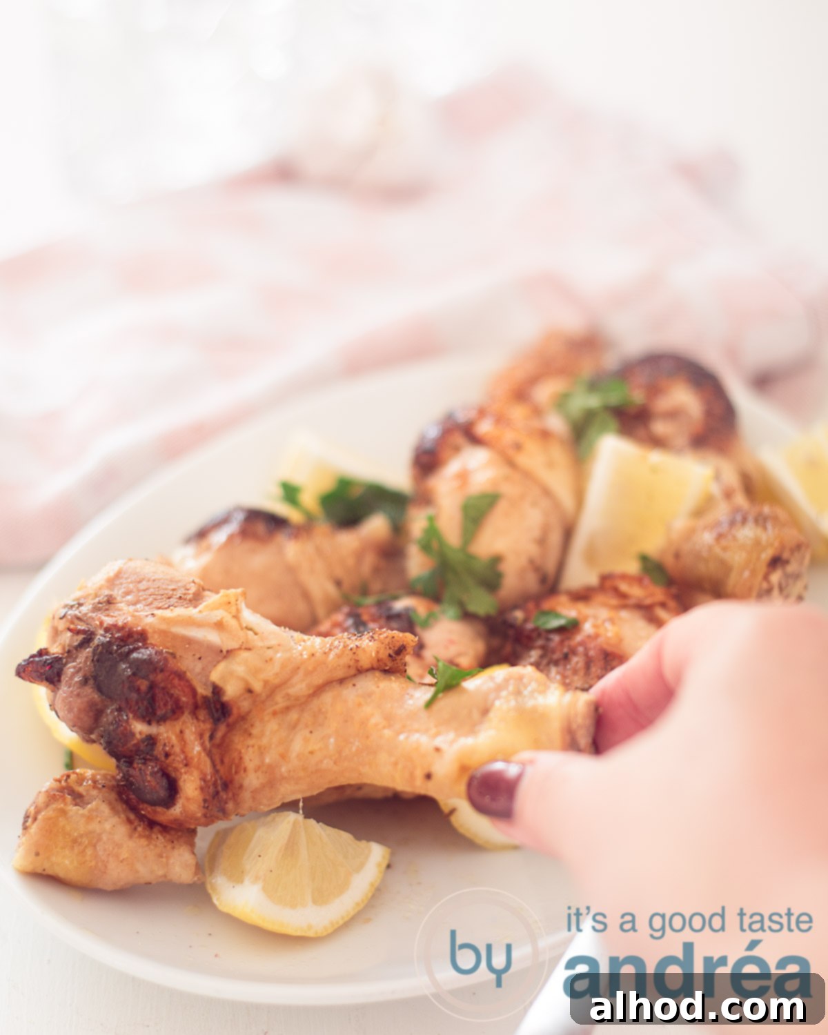 A white long plate with golden-brown chicken drumsticks, fresh lemon wedges, and vibrant parsley garnish. A hand reaches for a drumstick, suggesting the dish is ready to be enjoyed. A pink and white checkered cloth subtly decorates the background.