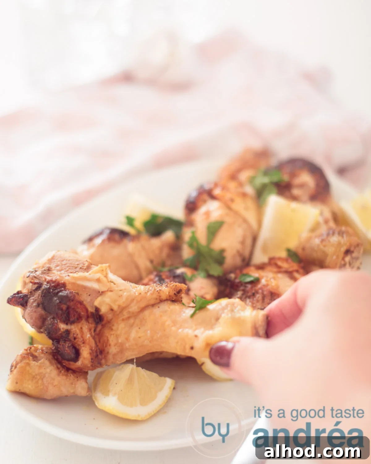 A white long plate with drumsticks, lemon parts and parsley. A hand taking a drumstick. A pink white cloth in the background