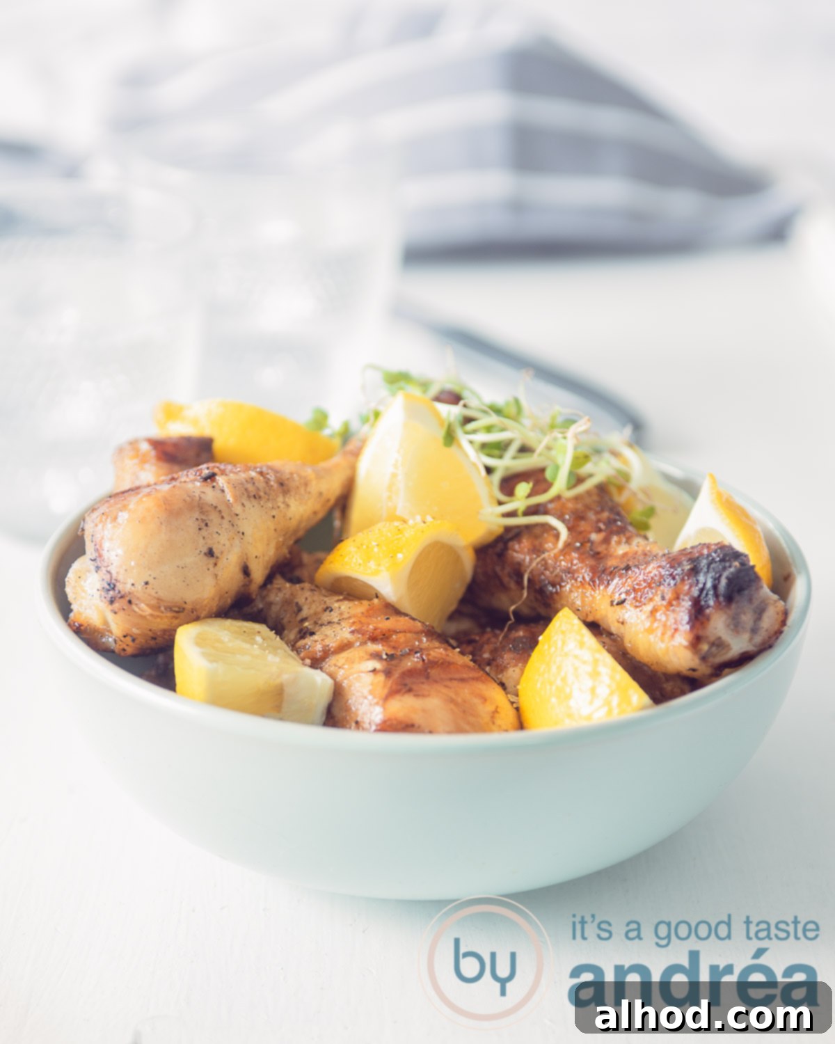 Close-up of golden-brown chicken drumsticks with fresh lemon and parsley in a blue bowl, set against a simple white background with a grey and white cloth.