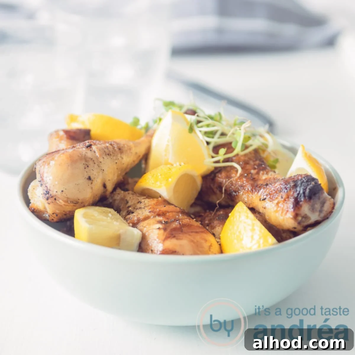 A square photo with a blue bowl filled with drumsticks and lemon wedges on a white background. In the background a gray and white striped tea towel.