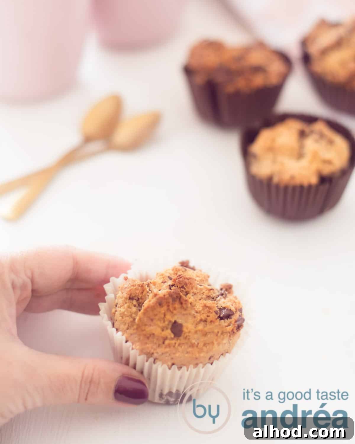 A hand picks up a chocolate and coconut muffin in a white paper tray. In the background three muffins in a black mold, to the left of that 2 spoons and two pink coffee mugs. Everything on a white background